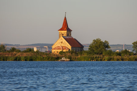 The Church of St. Linhart, a Romanesque-Gothic building with a characteristic red roof and tower, dominates the center of the photograph. The church stands on a small wooded island that forms an embankment in the middle of the NovomlÃ½nskÃ¡ reservoir on the PÃ¡lava River (South Moravia, Czech Republic)の写真素材