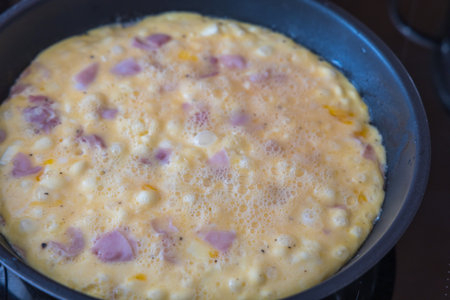 Close-up shot of an egg mixture with ham and onion pieces cooking in a black non-stick frying pan....