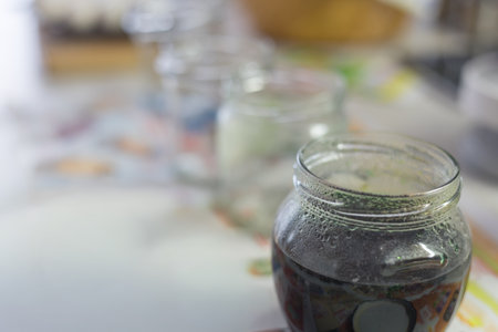 A row of jars with a dark liquid, Easter egg dye, on a light table. The foreground focuses on one jar, which is dewy. The blurred background suggests home preparation for Easter.の写真素材