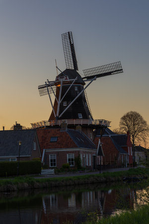 Traditional windmill De Vlijt in Bedum, Netherlands, at dusk. The illuminated mill stands above brick houses and reflects in the calm canal water.の写真素材