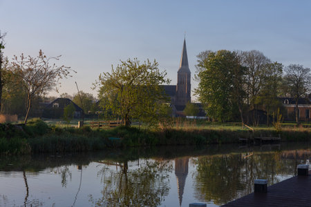 Morning view of the Walfriduskerk church tower in Bedum across a calm canal. Typical Dutch landscape with reflections and spring greenery.の写真素材