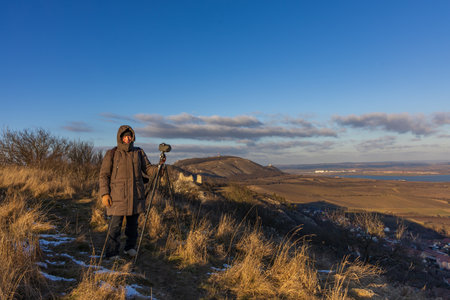 Portrait of a female photographer standing by a tripod in the winter Palava landscape at sunset. View of snowy hills and reservoirsのeditorial素材