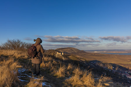 A female photographer with a backpack and tripod takes photos of the winter Palava panorama at sunset. View of snowy hills and reservoirsのeditorial素材