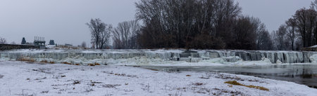 Breathtaking shots of a frozen dam on the Jihlava River. Massive ice formations, icicles, and frozen waterfalls covering the weir structure. The contrast between flowing water and thick ice creates a dramatic winter landscape under a sunset skyの写真素材