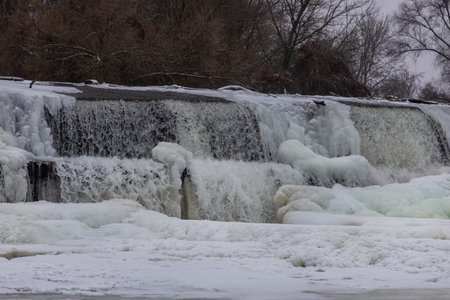 Breathtaking shots of a frozen dam on the Jihlava River. Massive ice formations, icicles, and frozen waterfalls covering the weir structure. The contrast between flowing water and thick ice creates a dramatic winter landscape under a sunset skyの写真素材
