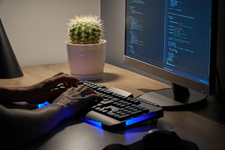 Teenage man (student) working late night in front of computer screen. He is programming in python language using keyboard with blue light on his deskの写真素材