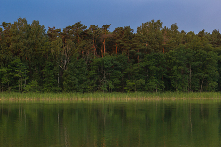 A reflection of forest and shore in a calm lake under a dynamic sky.の写真素材