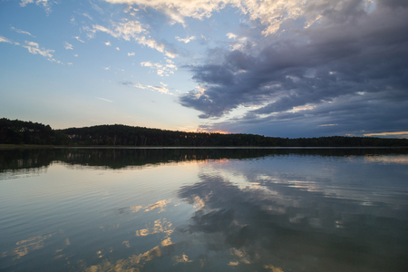 A reflection of forest and shore in a calm lake under a dynamic sky.の写真素材