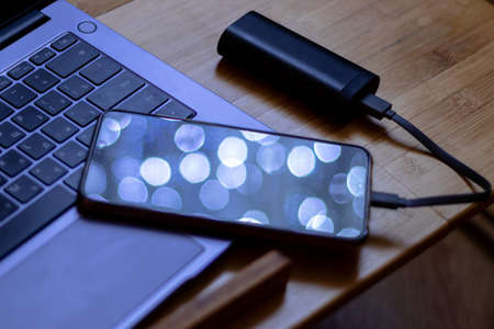 Phone is charged by the power bank. The phone is lying next to the laptop and is being charged by the power bank in a dark room, lights are reflected on the display.の写真素材