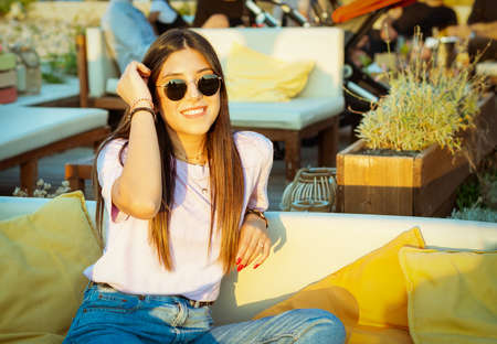 beautiful smiling caucasian girl with brunette hair and sunglasses sitting in a lounge bar at sunset...