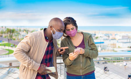 happy interracial couple wearing face mask interacting with smatphones on the seaside in a autumn...