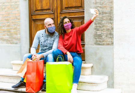 happy couple of a bearded bolt man and a curly caucasian woman wearing protective face mask carrying shopping bags sitting on stairs in the city center during black friday sales.girl and a boy taking a selfie .concept about consumerism and new normal lifestyleの写真素材