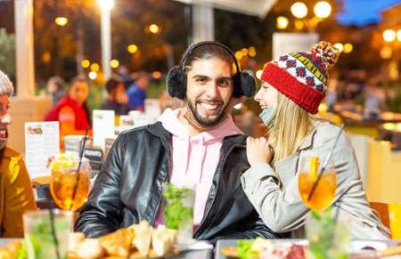 happy couple of a caucasian man wearing funny earmuffs and a blonde smiling woman with a winter hat.girl and a boy celebrating festivities wearing protective face mask in a bar.concept about togetherness and joy with friendsの写真素材