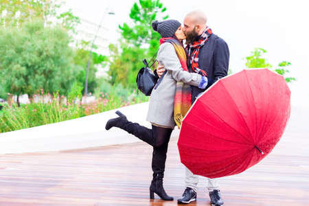 group of a bearded bolt man holding an umbrella and a caucasian brunette woman wearing warm winter clothes. couple of a boy and a girl kissing under the rain.concept about love, romance, passion and togethernessの写真素材