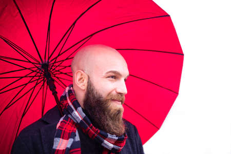 bolt bearded man wearing a scarf and warm winter clothes holding a red umbrella as a background.smiling young adult on a white scenario in a studio shooting.concept about joy, happiness and natural beautyの写真素材