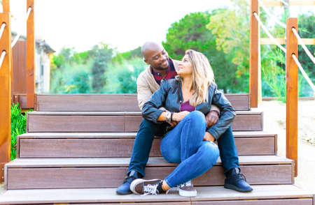 happy group of a multiethnic bolt man and a caucasian woman looking at each other sitting on stairs in a seaside park during autumn holidays.smiling interracial couple of a boy and a girl chilling and relaxing outdoors.concept about love and friendshipの写真素材