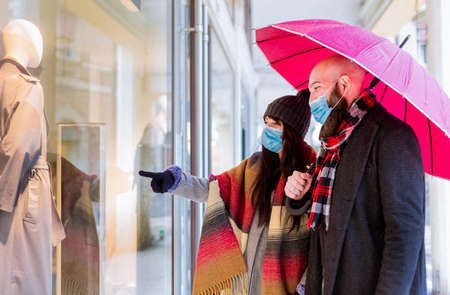 Beautiful couple with an umbrella and winter clothes is talking and smiling while doing shopping in the city in a rainy day.side view of a couple walking past a shop window and looking in for sales after lockdown reopening.new nomal lifestyleの写真素材
