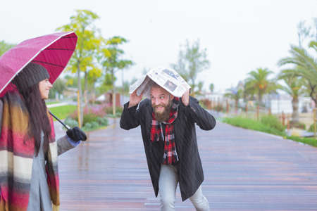 Cheerful pretty girl holding umbrella while her boyfriends runs under the rain under a newspaper outside. She is turning and looking at him with true delight and sincere smile and love. Double of a bearded bald man and a beautiful woman in a strorm.の写真素材