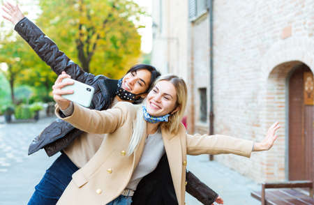 Young people taking selfie on city street wearing face protective masks.Coronavirus prevention, travel safe, technology trends and autumn vacation concept.couple of funny women during new normal lifestyleの写真素材