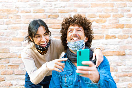 Cheerful best friends viewing funny photos in social networks via smartphone standing together on a street wall background.happy male and female watching funny online video content outdoors after lockdown reopening.new normal lifestyleの写真素材