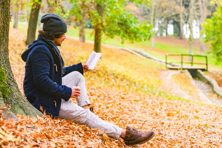 young curly latino man sitting on the ground in a park drinking a coffee to go and reading a book during autumn looking at a river.red and orange mood.Beautiful landscape with yellow trees and Colorful foliage in the park.の写真素材