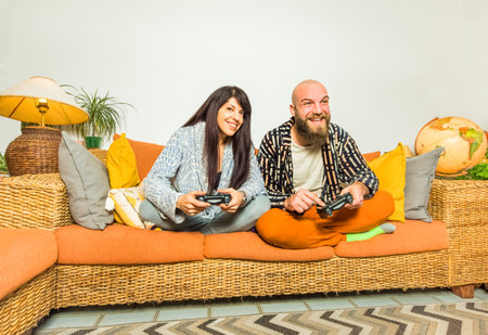 happy couple of a hipster bald man and caucasian brunette woman holding two console joypad sitting comfortable on a couch for gaming evening.fun, togetherness, joy, tech and youth concept. home lifestyleの写真素材