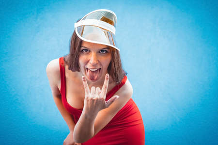 close up portrait of one young cheeky beautiful woman standing on colorful background wall.attractive charming girl showing tongue, heavy metal sign symbol.female joking wearing red evil dress and hat.の写真素材
