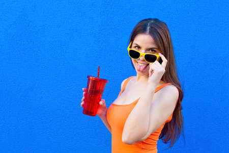 Young beautiful woman with sunglasses standing on background wall drinking from reusable bottle canteen.happy caucasian girl wearing colorful summer clothes smiling at camera making funny faces.joy.の写真素材