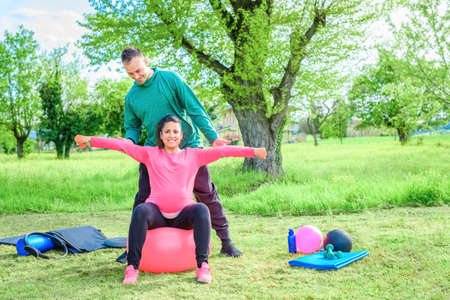 couple of a smiling personal trainer and a beautiful pregnant woman doing workout exercise outdoor in a park.girl lifting weight sitting on gym ball. fit, training, fitness, health and friendship conceptの写真素材