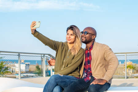 happy group of a multiethnic bolt man and a caucasian woman taking a selfie sitting on a bench in a park during spring holidays.smiling interracial couple of a boy and a girl taking a self portrait photo outdoors.sunlight hitting on themの写真素材