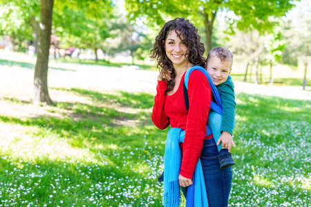 happy young mom carries a child on her back, tied up with a colorful traditional piece of fabric in a public park garden. mother and son in a baby wrap sling chilling outdoor.joy, family and togethernessの写真素材