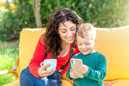 happy mom and son chilling outdoor sitting on couch in public park garden watching funny videos on smartphone.smiling woman and child interacting online on apps. tech, social media influencer conceptの写真素材