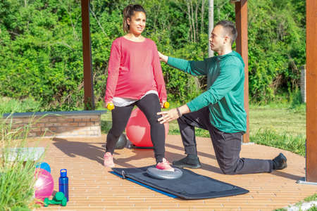 Young pregnant woman training arms with weights lifting standing on a mat followed by her coach. personal trainer train a girl outdoor in park. fitness, fit, maternity, healthy lifestyle concept.の写真素材