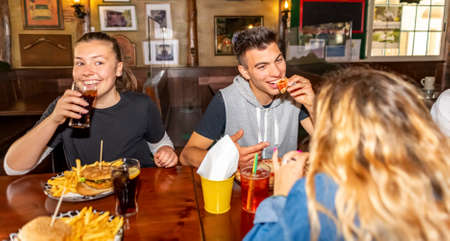 Group of millennials friends eating at fast food indoors in irish pub restaurant. Happy people partying and eating together. Young active teenagers with burgers, pizza and drinks having fun dining.の写真素材