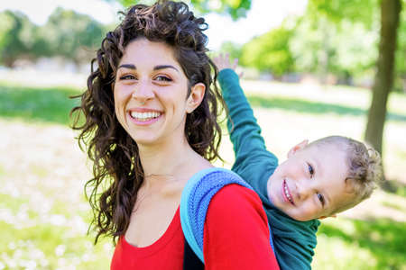 close up portrait of a happy young mom carries a child on her back, tied up with a colorful traditional piece of fabric in a public park garden. mother and son in a baby wrap sling chilling outdoor.の写真素材