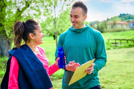 couple of personal trainer and a young student woman pregnant discussing training schedule together smiling outdoor. fitness and healthy lifestyle concept about workout, fit, friendship in parkの写真素材