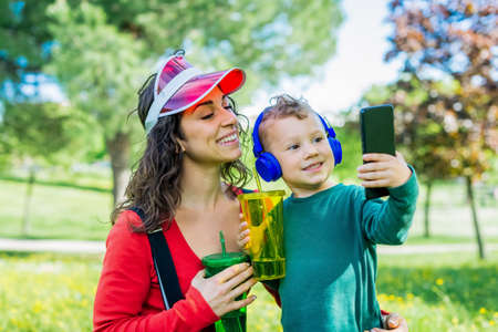 happy mother and little kid wearing headphones interacting with technology listening to music outdoors in a park spring. mom and child time chilling in a garden drinking and making selfie. joy conceptの写真素材