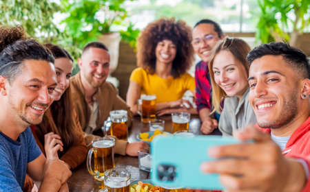 diverse group of friends making a selfie celebrating happy hour with beers and food in a bar restaurant. multiethnic people having fun in a party together smiling at the camera. self portrait conceptの写真素材