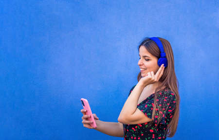 happy wonderful woman wearing headphones standing against a blue background wall. Smiling girl listening to music holding a smartphone wearing summer clothes. tech, joy, relax and chilling concept.の写真素材
