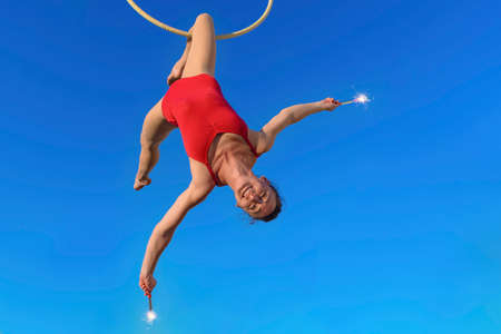 acrobat athletic, young graceful gymnast performing aerial exercise in the air ring outdoors isolated on background holding fountain sparklers. woman in red suit performs circus artist dancing on hoopの写真素材