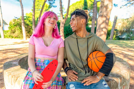 alternative diverse couple hanging out together chilling and smiling sitting in a park holding a...