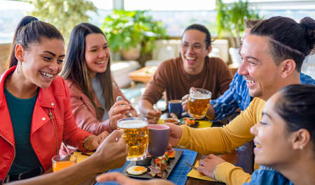multiethnic group of friends sitting on a table in a bar restaurant making a toast with beers and drinks. diverse people celerating brunch together enjoying happy holidays. lifestyle and joy conceptの写真素材