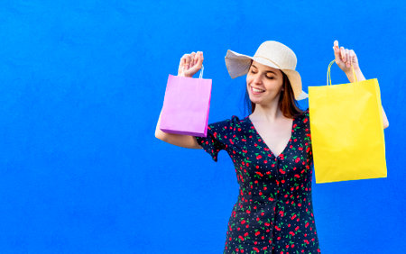 young happy woman wearing summer clothes holding shopping bags isolated on a blue background wall. girl shop gifts on black friday celebrating sales. retail, purchase, fun, joy, and lifestyle conceptの写真素材