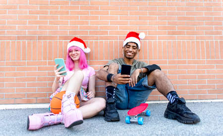 merry christmas! happy diverse couple of friends wearing xmas santa hat interacting with smartphone sitting against background wall together on winter warm festivities outdoor. summer holiday conceptの写真素材