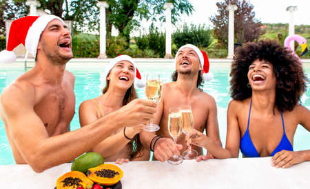merry christmas! happy diverse group of friends wearing xmas santa hat having champagne wine drinks at swimming pool party making a toast celebrating winter warm festivities outdoors. summer holidaysの写真素材