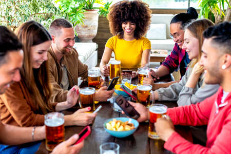 young multiethnic group of friends holding smartphone on social media having fun together drinking happy hour beer at bar restaurant sitting on table indoor. group of diverse people cheering together.の写真素材