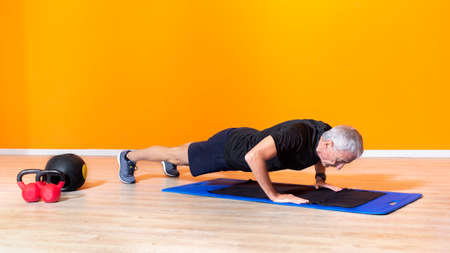 focused senior fit man doing push up workout exercise laying at the gym isolated on orange background. mature people fitness training indoor. elderly person recovery healthy lifestyle conceptの写真素材