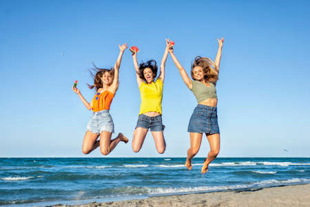 three happy girls on jumping motion at the sand beach outdoors in summer celebrating hot vacation days. happy funny friends having fun on warm seaside holidays joy, carefree, genz and party conceptの写真素材