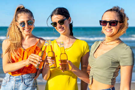 three happy women toasting beers at the beach standing on sand outdoor celebrating summer vacation days looking at camera. friends on happy hour seaside sunset holiday. freedom, genz and party conceptの写真素材