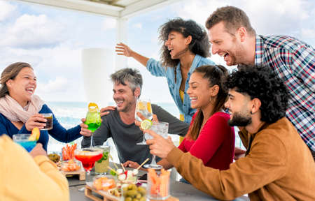 interracial group of friends at rooftop terrace bar at the beach having food and drinks celebrating happy hour on summer weekend. diverse people together enjoying holidays. fun and lifestyle conceptの写真素材